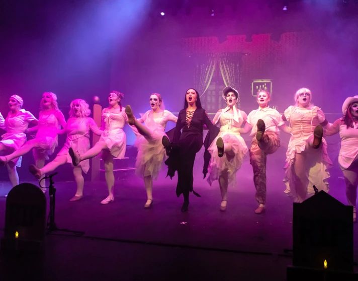 Stage performance featuring a line of dancers in ghostly white outfits, led by a character in dark clothing, all kicking in perfect unison. Stage performance featuring a line of dancers in ghostly white outfits, led by a character in dark clothing, all kicking in perfect unison.